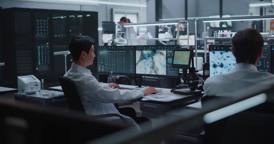 Asian Male Scientist Logs Microscopy Images and Cellular Datasets on Monitors While Lab Team Processes Samples in Biotech Center for Drug Development, Diagnosis and Disease Therapy Research