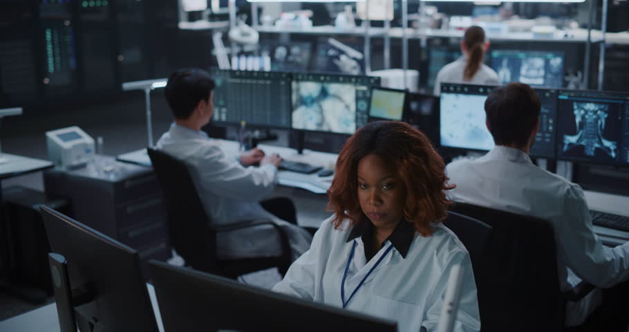 Portrait of Black Female Scientist in White Lab Coat Works Focused at Computer in Modern Medical Research Facility as Colleagues Use Ai to Analyze Brain Scan Data for Drug Development in Background.