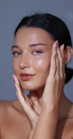 Close-up of beautiful young woman gently touching face with hands in vertical format. Soft lighting highlights glowing skin. Perfect for beauty brand commercials and skincare ads.