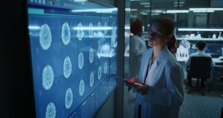 Female Scientist Examines MRI Brain Scans on a Large Touchscreen Display While Holding a Tablet in a Modern Research Laboratory, Analyzing Neurological Data for Drug Development and Disease Treatment.