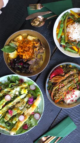 Friends enjoying a vibrant and healthy lunch together at a restaurant, eating delicious asian fusion dishes like ramen, curry, and fresh salads with chopsticks from a dark wooden table