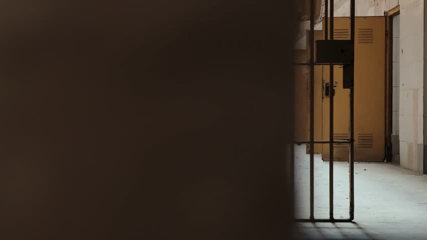 View through prison bars inside an old abandoned jail corridor with locked gates and dim natural light. Atmospheric detention facility interior, crime, punishment and justice system concept. High