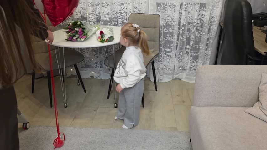 Cute little toddler girl holding a red heart balloon ribbon at home.