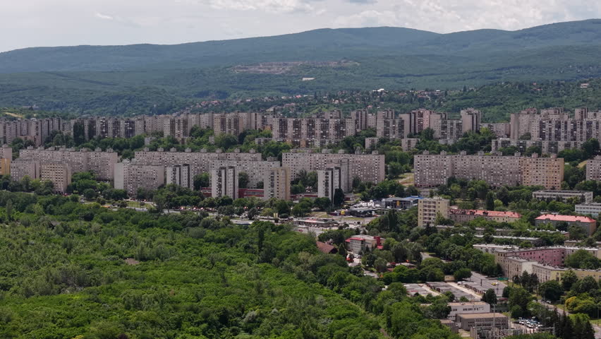 A sprawling socialist-era housing estate in Hungary, nestled against a backdrop of lush green hills under a cloudy sky.