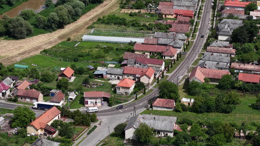 An aerial view reveals a charming Hungarian village, with houses lining a main road, green fields, and a river, depicting tranquil rural life.