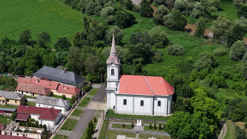 An aerial view showcases a white church with a red roof and steeple, surrounded by buildings and lush green nature in Hungary.