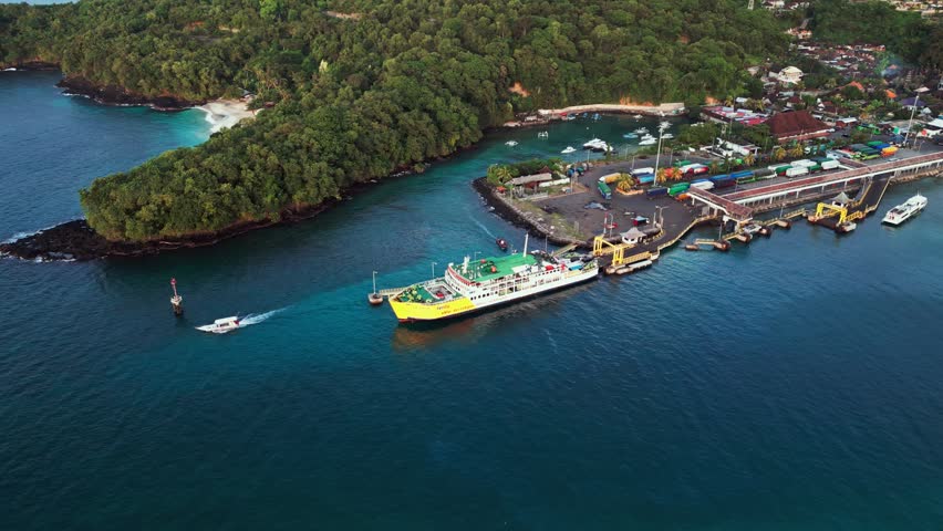 Passenger ferry docking at a tropical island port harbor along a lush forest coastline with a speed boat cruising the deep blue ocean water, aerial drone footage