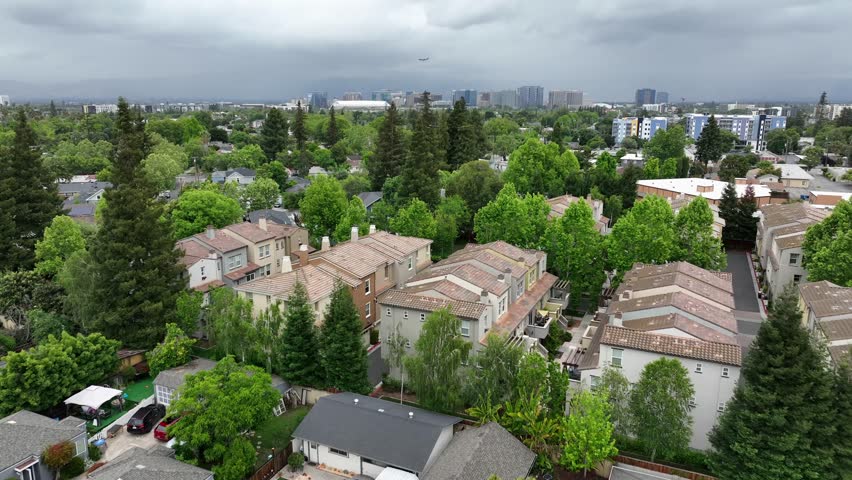 Storm clouds gather above red-tile townhome rooftops and trees, dense residential community giving way to San Jose skyline on distant horizon. San Jose, California, USA. Aerial drone flyover shot