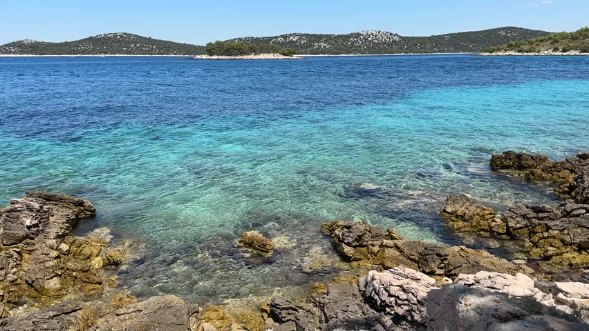 Footage shows a clear blue sea near a rocky coast in Croatia. Green hills are visible in the distance under bright sunlight. The water ripples gently as waves touch the rocky shore.