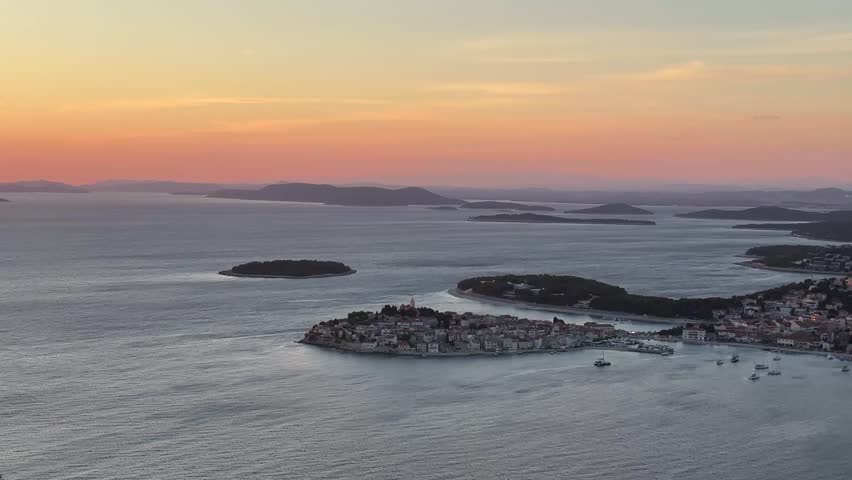 A view shows the sunset lighting up the sky over the islands and a small town along the coast of Croatia. Small boats are visible in the water below the colorful sky.