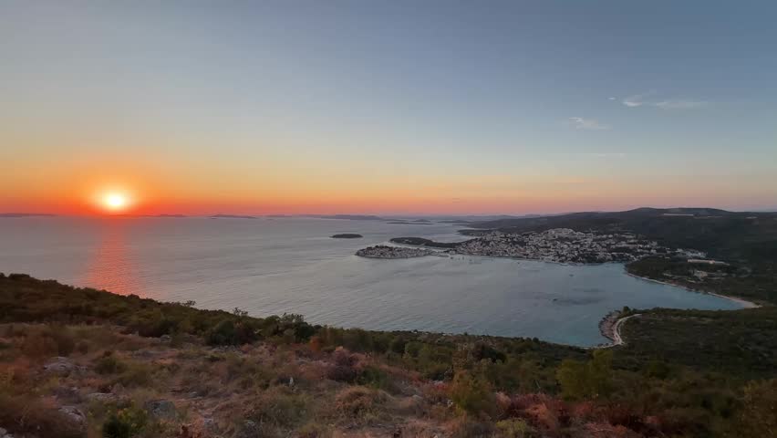 The sun sets over the water and town, filling the sky with bright colors. The coastline shows hills and the quiet town along the shore. This happens in the evening in Croatia.