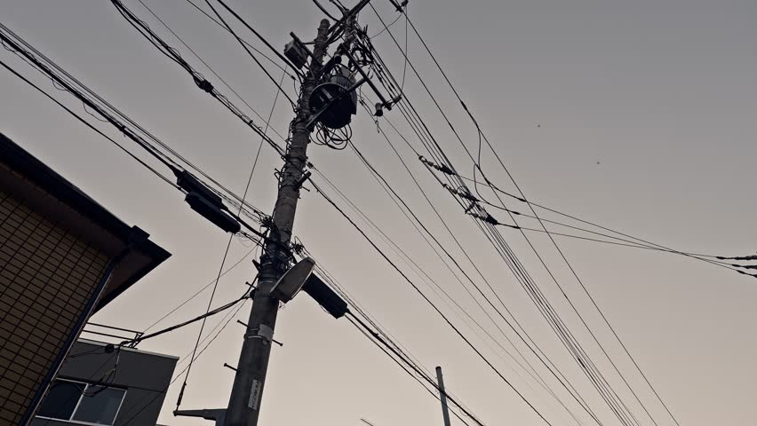 A concrete pole bearing a transformer and branching cable connections rises beside a brick building corner. Multiple wires radiate outward against a pale gray dusk sky.