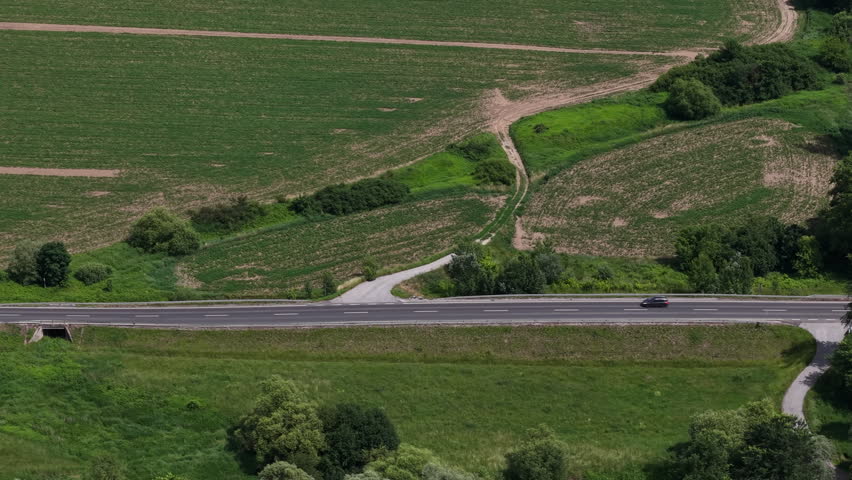 An aerial view shows a lone car driving along a highway through expansive green agricultural fields in rural Hungary.