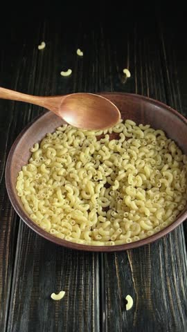 Wooden spoon lifts elbow macaroni from a brown bowl, showcasing the pasta