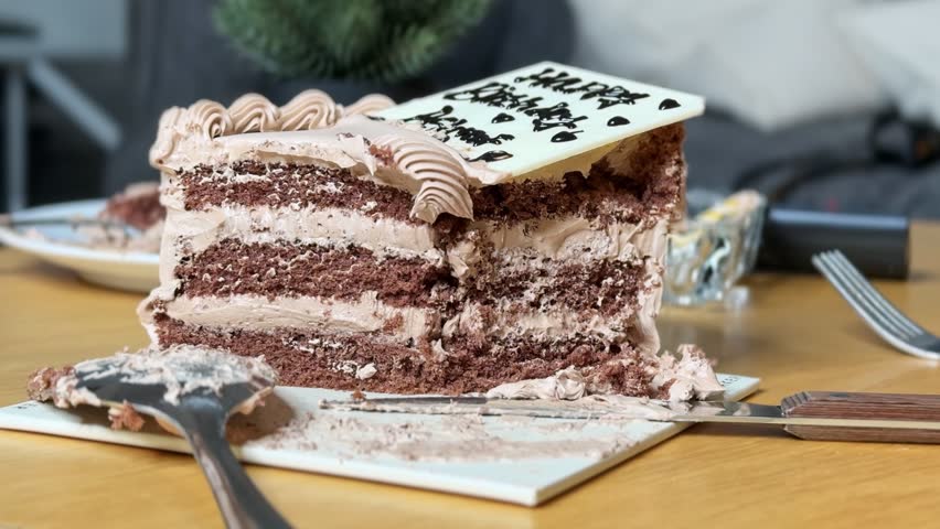 Close-up of a partially eaten layered chocolate birthday cake with a white plaque resting on a messy wooden table. A fork and spoon sit nearby in this modern indoor living room setting during daytime.