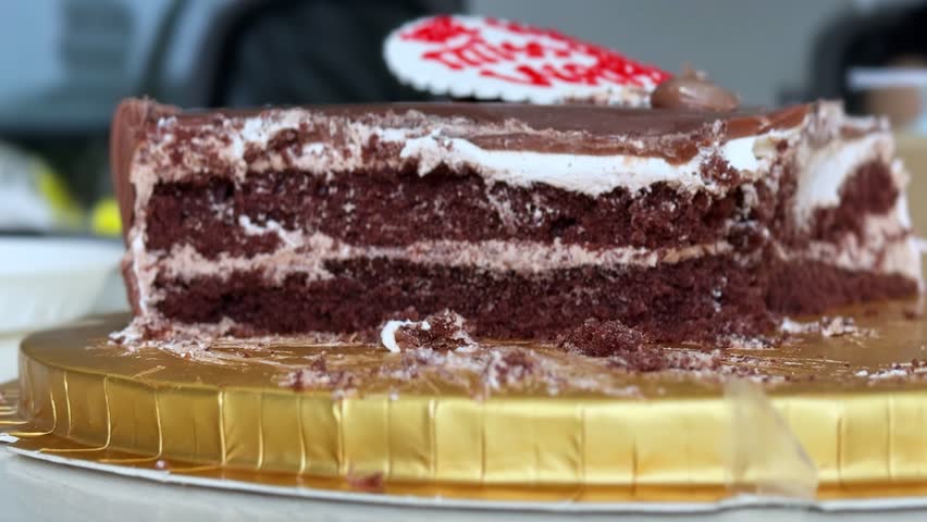 a partially eaten layered chocolate cake resting on a gold cardboard tray. Slices have been removed from the sweet dessert, leaving crumbs on a white table in a bright indoor setting.