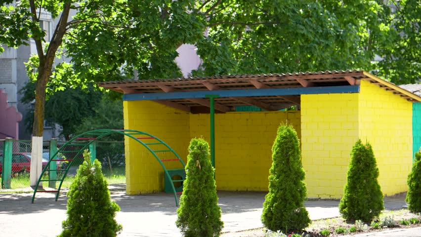 A bright yellow brick playground pavilion with a brown roof stands behind a neat row of small green thuja trees on a sunny summer day.