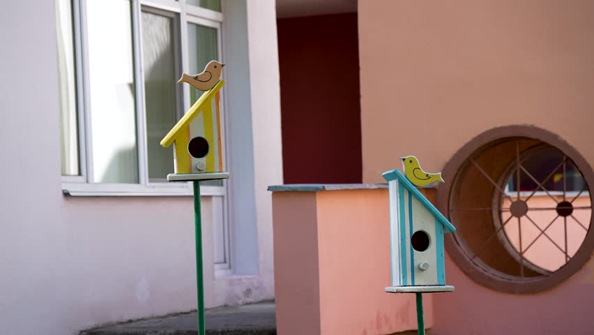 Two charming, handcrafted wooden birdhouses painted in pastel yellow and blue tones stand on green poles as decorative installation pieces near a building courtyard.