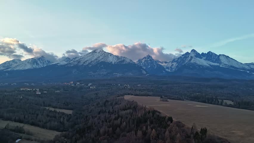 In the High Tatras of Slovakia, snow-capped mountain peaks rise above forest and a mountain village in a drone hyperlapse panoramic winter view.