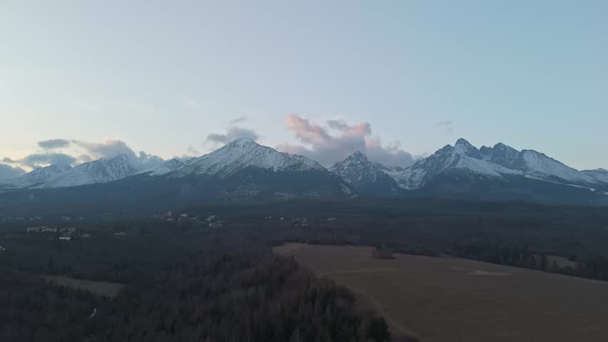 In Nová Lesná, Slovakia, snow-capped peaks of the High Tatras rise above coniferous forest and rural meadows, viewed in a wide drone panorama under a broad sky with scattered clouds.