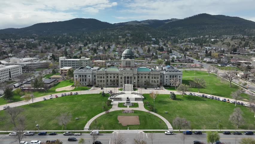 Circular pathway and Montana-shaped garden bed front Capitol building, curved walkways framing lawn beneath forested mountains and partly cloudy sky. Helena, Montana, USA. Aerial drone approaching.