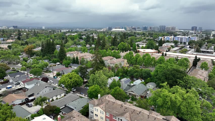 Storm clouds loom over San Jose neighborhood, single-family homes and townhomes nestled among trees with downtown skyline rising in distance. San Jose, California, USA. Aerial drone flyover shot