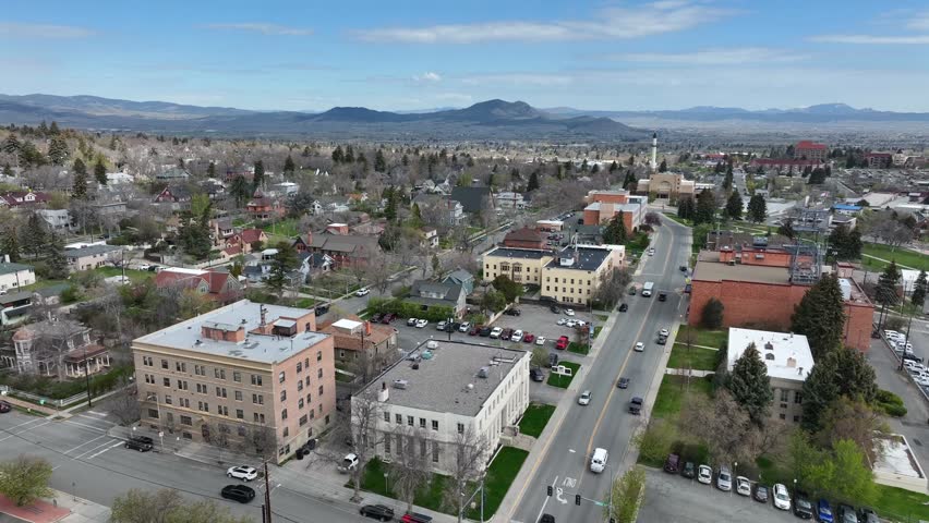 Classical brick and stone buildings line street intersection, distant minaret tower rising above rooftops beneath mountain range and parking lots. Helena, Montana, USA. Aerial drone descend shot