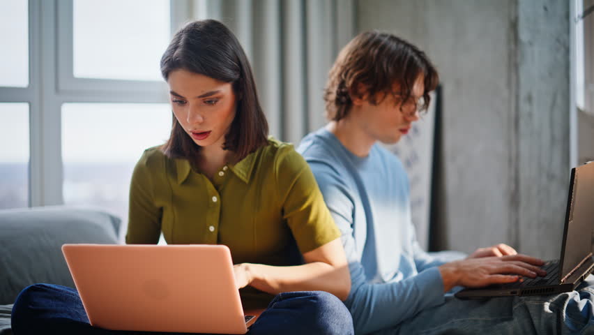 Enthusiastic pair creating startup at home sitting bed in cozy atmosphere closeup. Smiling two people looking each other working online using laptops. Cheerful students studying remote make homework 