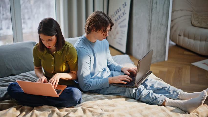 Girlfriend boyfriend browsing laptops at soft bed closeup. Happy man positive woman typing keyboards looking each other with smile. Young couple working computers together at light apartment interior 