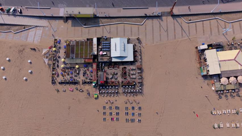 A magnificent top-down aerial photograph capturing the detailed layout of a seasonal beach club and restaurant pavilion on the wide sandy coast of Scheveningen in The Hague. The center of the frame features a large modular beach structure showcasing a mix of architectural elements, including flat eco-friendly green living roofs, solar panel installations, and light-colored corrugated roofing. Surrounding the pavilion are expansive wooden outdoor dining terraces meticulously arranged with rows of small dining tables, umbrellas, and vibrant colorful chairs. Below the terrace, neat geometric rows of empty sun loungers and deck chairs are laid out directly on the smooth, tan sand. The top edge of the frame captures a section of the paved concrete pedestrian seaside promenade boulevard, showcasing a clean, modern coastal layout from a perfect bird