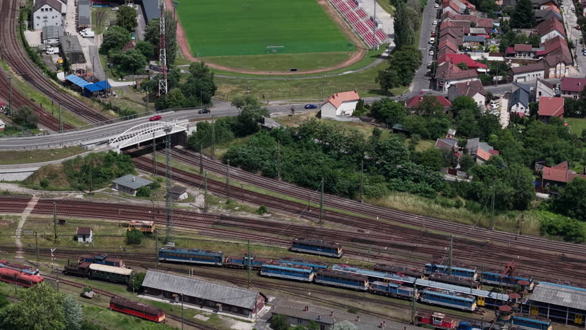An expansive aerial view shows multiple trains at a railway junction near a sports stadium and neighborhood in Hungary.