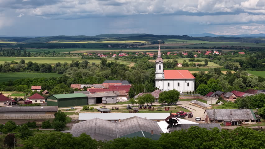 An aerial view of a traditional Hungarian village with a church steeple overlooking vast green farmland and rolling hills.