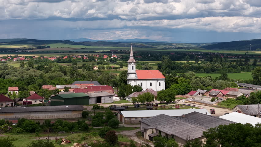 A scenic aerial view captures a traditional church in a small Hungarian village surrounded by green fields and rolling hills.
