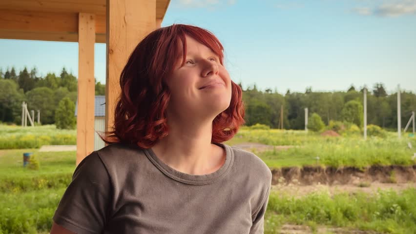 A woman is looking upwards and smiling in a happy outdoor setting under a wooden structure with a green field in the background. The scene is captured from a slightly elevated angle.