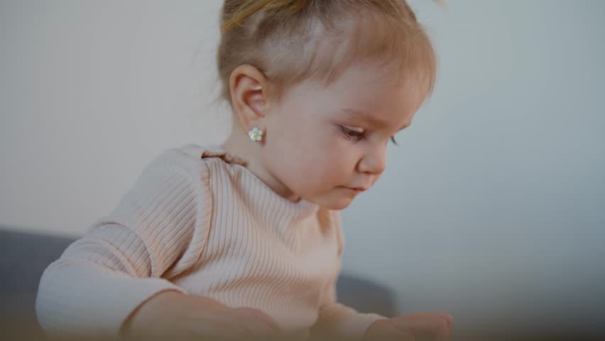 Young girl with light hair tied in pigtails sits on a couch. She wears a light-colored sweater and pearl earrings. Her gaze is focused downward, showing concentration. She moves her hands gently