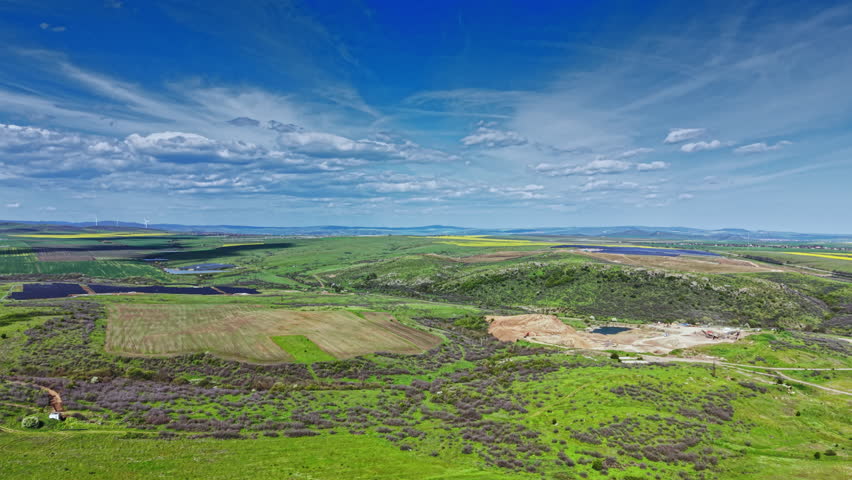 In the late afternoon, green fields stretch across the rural landscape with patches of cultivated land. Distant hills and a blue sky with clouds create a vast view.