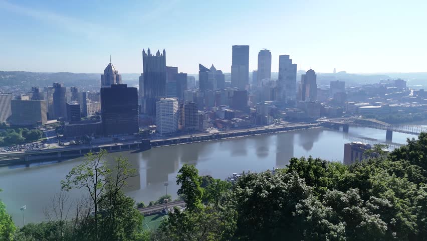 Scenic wide drone viewpoint of the Pittsburgh skyline and skyscrapers from a high green hillside overlooking the river