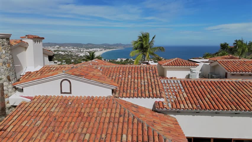 Drone ascends over a hilltop rooftop in San Jose del Cabo on a bright sunny day, revealing the city, main highway, and stunning Baja coastline stretching into the distance.
