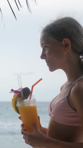 Vertical video. Teenage girl drinking a fresh mango smoothie through a straw with a pineapple slice garnish on a sunny tropical beach with the sea visible in the background. Summer refreshment