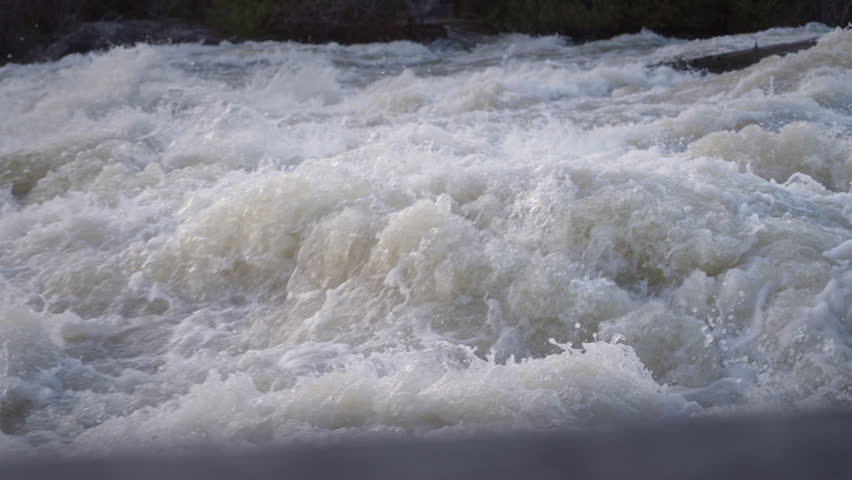 Spring runoff rushes violently downstream at Sauble Falls, Ontario. The slow motion capture reveals the complex fluid dynamics of churning white foam and splashing water droplets.