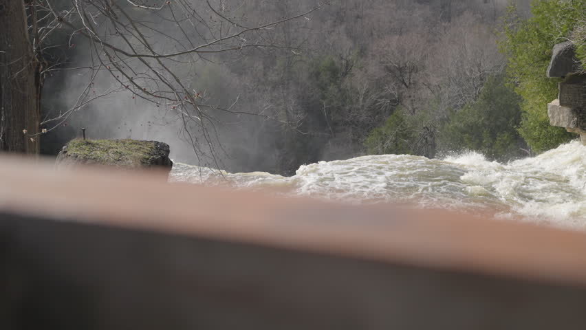 Rushing river water flows rapidly over the limestone crest of a powerful waterfall. Splashing droplets and thick mist rise from the gorge below in slow motion detail.