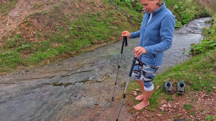 Woman crossing mountain stream side view in Georgia, walking barefoot through cold water with trekking poles on rural hiking route for travel, river safety, trekking tourism, and expedition footage