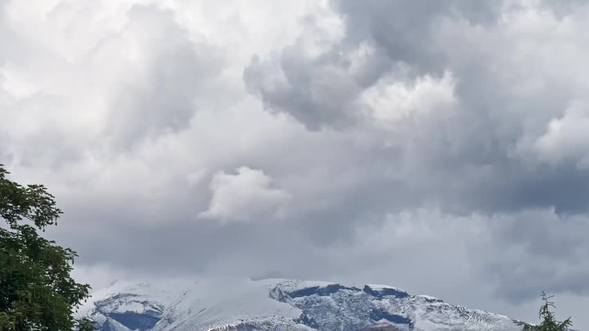 Heavy storm clouds hang over snow-capped alpine peaks, with a dark tree silhouette framing a dramatic mountain weather scene.