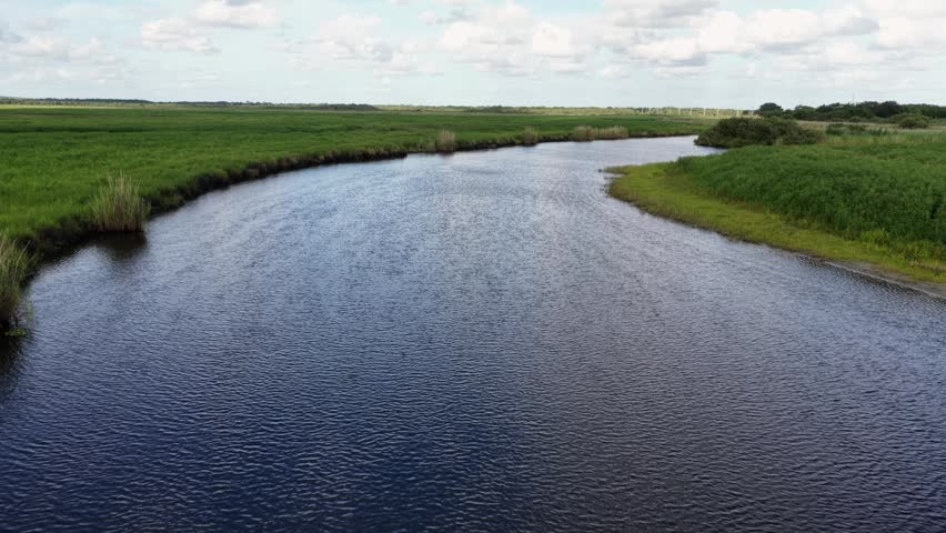 Winding river channel curves through vast Everglades marshland, dense green grasses, rippling blue water, scattered clouds, remote flat wetland, Florida, United States. Aerial drone flyover shot