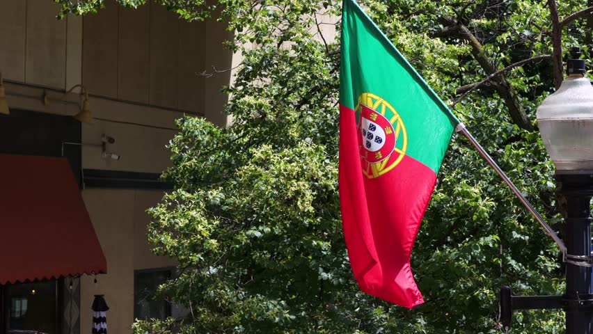 Portuguese flag waves near red restaurant awning, sunlit green tree canopy, security camera, storefront windows, festive soccer World Cup 2026 spirit, small American town, United States. Aerial close