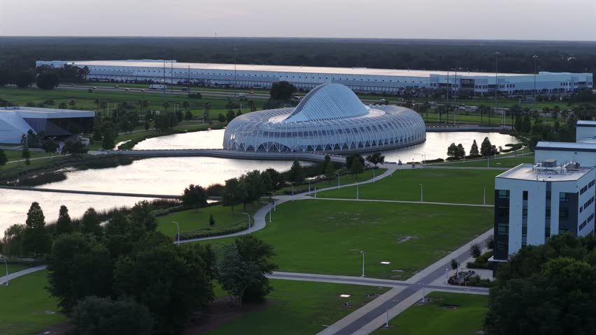 Futuristic saucer-shaped building anchors Florida Polytechnic University, reflective lake, green lawns, walking paths, evening light, Lakeland, Florida, United States. Aerial drone wide shot