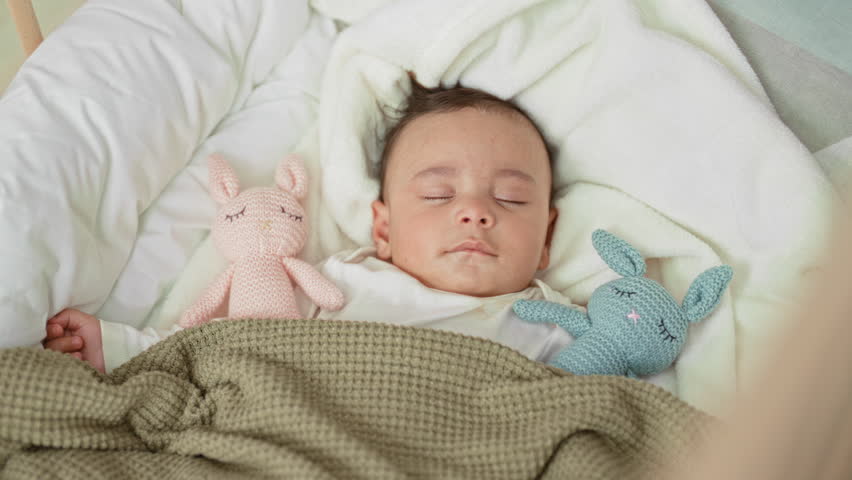 Baby sleeping in cradle with visible left hand and two knitted bunny toys on a soft blanket in a nursery; calm comfort.