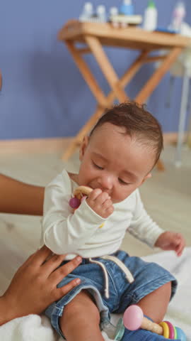 Baby boy sits on blanket and holds teether to mouth, chewing with hand near lips inside a building; curious comfort.