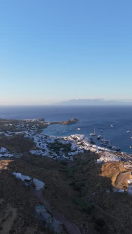 Mykonos island in Greece revealing white washed houses on coastal hills with yachts in Aegean Sea harbor and distant islands under clear blue sky