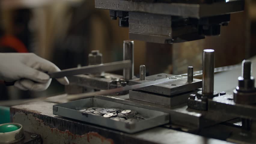 Worker adjusts metal mold with gloved hands. Machine hovers above coins ready for pressing. Tooling shows industrial precision in a factory. Scene captures manufacturing focus and craftsmanship